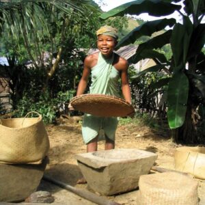 Girl holding broad, shallow woven basket with large wooden mortar and pestle on ground in front of her