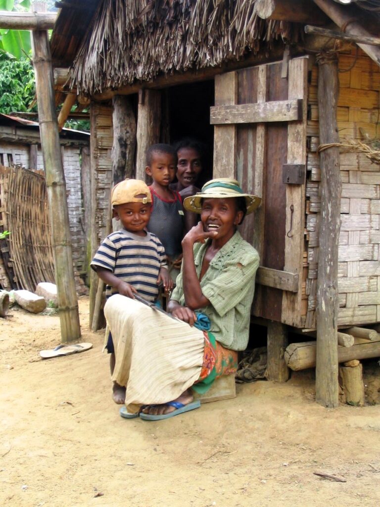 A woman in a hat sits with a small boy at the doorway to a small house made of bamboo and palm fronds
