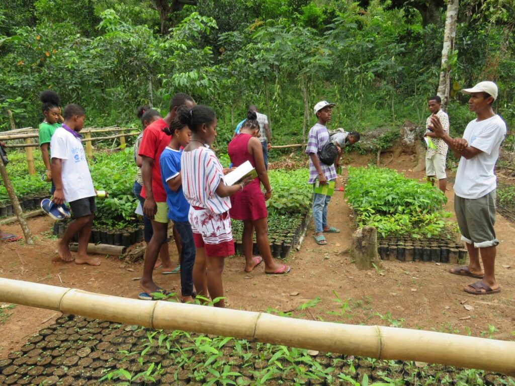 A man is talking to 11 young students in a tree nursery. The tree nursery is set in a clearing in a forest. It has hundreds of small pots of tree seedlings.