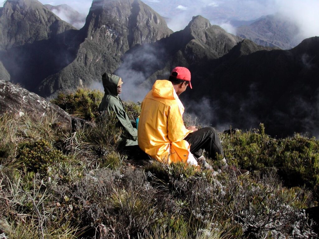 Two people wearing raincoats sit on a grassy hillside with steep, cliffy mountains shrouded in clouds in the distance.