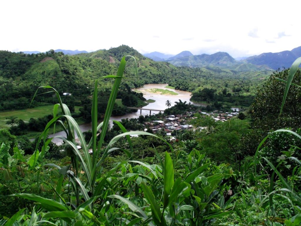 Looking down from high on a hill to a small village with many tiny houses on the banks of a large, meandering river. Green hills and rice fields lie on either side of the village and river.