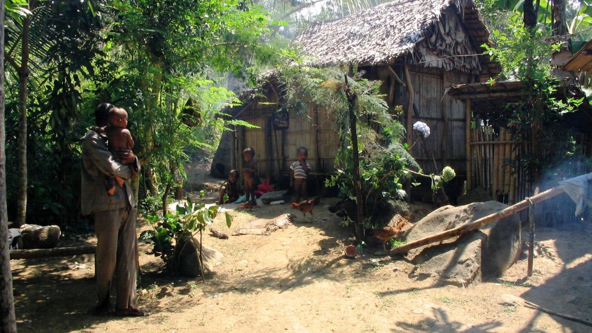 A man holding a baby and several small children stand around a small, thatched hut deep in the forest.