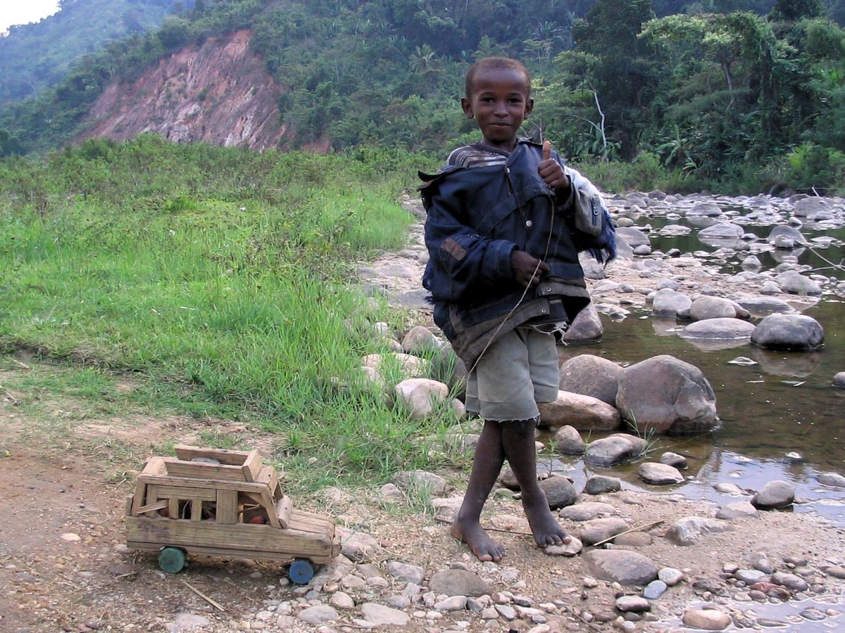 Small boy, smiling, standing at river's edge, dressed in raggedy blue jacket, pulling small wooden toy car by a string. Green, forested hills in the background.
